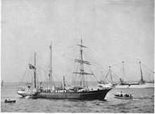  A three-masted ship with sails furled, short funnel amidships, flag flying from the stern on left of picture. Two small boats are close by, and a larger vessel decked with bunting is visible in the background.