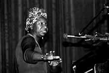Head and torso of black woman at a piano in front of a microphone wearing a flowered kerchief in her hair