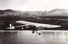 Single-engined high-wing light plane flying over water, with jungle terrain in background