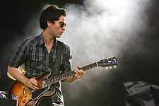 Kelly Jones with gelled up hair on stage, wearing sunglasses, playing a semi-hollow Gibson electric guitar while singing.