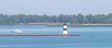 A pier with a square, black and white-striped lighthouse at the end. A small motorboat is paralleling the pier, heading away from the lighthouse.