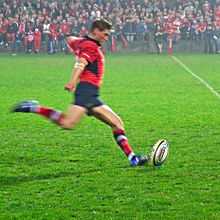 A man in a red shirt is running forward to kick a rugby ball placed on a tee.