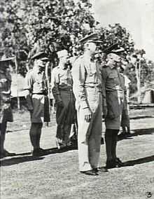 Full-length, three-quarter-angle, outdoor portrait of half a dozen or so men in light tropical military uniforms with headgear, standing to attention. Two of the men are in the foreground and the rest in a row behind them.