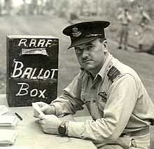 Man in military uniform with peaked cap seated at a desk beside a box marked "RAAF Ballot Box"