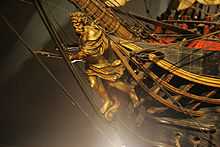 An old-gold colored figurehead, still attached to the brown wooden ship, of a crowned, bearded man with a double-handed spear. He is covered on his forward arm and just below his waist by flowing cloth. With one foot back towards the ship and one foot forward on gold scroll-work at the bow, he shown starting a charge forward, aiming his spear ahead.