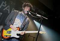 A male vocalist plays electric guitar in front of a mic stand. The stage is lit by white lights and its background is mostly black.