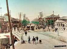 Street scene: People walking over a bridge, with low-rise buildings, street cars, and utility poles leaning crookedly in the background