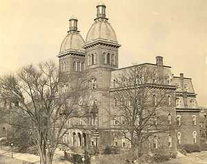 A 4 story building with two identical towers at the top surrounded by leafless trees