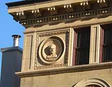 Upper corner of building, with dentils and brackets below roofline; medallion containing high-relief bust of Demosthenes