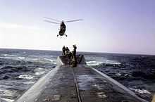 Surfaced nuclear submarine USS Triton with a six-man deck handling party standing on the raised housing located on the after end of the main deck while a U.S. Marine Corps Sikorsky UH-34 Seahorse helicopter hovering overhead.