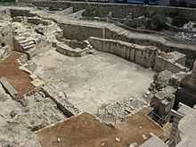 Looking down into excavated layers of ancient buildings with a large, irregularly shaped plastered pool at the bottom