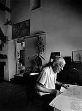 A black-and-white picture of a man seated at a table; an artist's studio is visible behind him