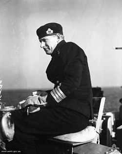 A man in naval uniform sitting on a seat on the bridge of a ship. He is wearing thick gloves and a cap, and is slightly side on to the camera. A body of water can be seen in the background.