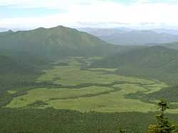 View from a mountain on a wide valley with light green areas bordered by dark green forest.