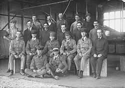 Group portrait of nineteen men, including six in military uniforms with peaked caps, in front of a biplane in a hangar