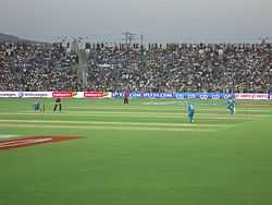 An image of a cricket field with the spectators in background. Players in blue outfit can be seen fielding, and a player in a red-blue outfit can be seen batting.