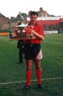A man with dark hair who is wearing a red top, navy blue shorts and red shorts. He is standing on a grass field, holding aloft a trophy.
