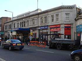 A pale two storey stone building, with gold coloured letters at the top reading "METROPOLITAN RAILWAY" and "PADDINGTON STATION", windows on the first floor. On the ground floor there are shops either side of an entrance with a canopy with rectanglar blue signs reading "PADDINGTON STATION". There are cars and a small truck in the road and people are walking on the pavement.
