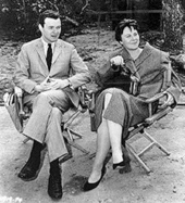 A black and white photograph of Alan J. Pakula seated next to Harper Lee in director's chairs watching the filming of To Kill a Mockingbird