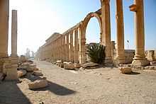 A crumbled array of stone pillars stand around an archway, lined along a dusty avenue.