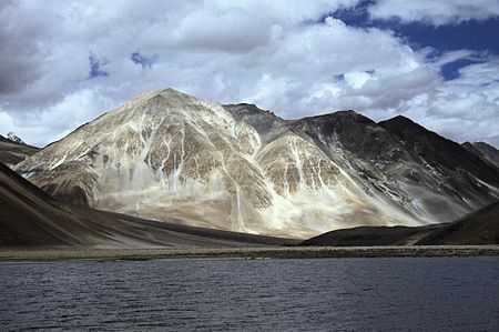 Daytime view of a large body of water standing before a prominent peak, which communicates with several others partly out of view and behind. A gravel beach at the far end of the lake gives way to steep slopes leading up to the peaks; The mountains lack trees. Patchy snowcover defines their recesses, and whitish vein-like streaks extend up from the base of the largest.