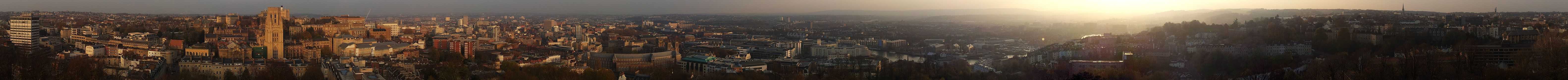 A photographic panorama of Bristol taken from the top of the Cabot tower. The picture shows an urban environment with densely packed offices and older buildings. Hills can be seen in the distance.