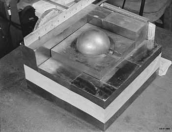 A stack of square metal plates with a side about 10 inches. In the 3-inch hole in the top plate there is a gray metal ball simulating Pu.
