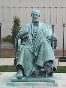 A green statue of a balding man with a monocle, seated and wearing long, flowing robes and holding a staff in his left hand