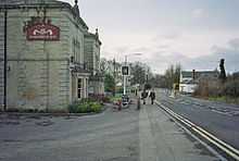 Gray stone building on the left with a pub sign outside it. A road is central to the picture with a white coloured building on the right.