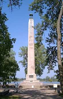 A tall, stone column at the end of a sidewalk, with trees on either side of the sidewalk.