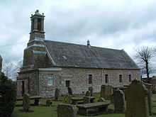 A grey stone chapel, with angular corners. An ornate bell tower is situated on the west of the building.