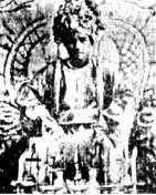 black and white photograph of a young cleanshaven man wearing an elaborate headdress and sitting on a throne. He is wearing robes and in front of him are some accoutrements on a table.