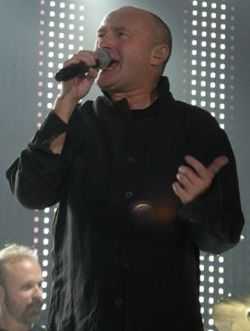 A man with a shaved head wearing black and singing into a microphone. The background contains dotted lighting on a stage.