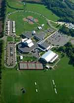 A clover leaf shaped building is seen from the air with tennis courts on one side and ball diamonds on the other.