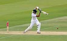 A man wearing a helmet and white clothes, in the motion of swinging a cricket bat.