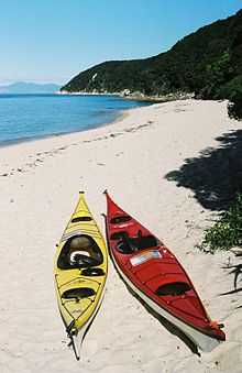 Photo showing clear blue water, a photographer or tourist capturing the water on a golden sanded beach and forested hills