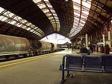  A railway station with curved platforms under an arched iron framed roof with roof-lights. A passenger train stands at the platform on the right and on the left passengers waiting for a train.