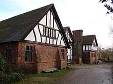 A house with three gabled bays seen from an angle.  The outer bays have lower storeys in brick with a timber-framed gable.  The middle gable is almost completely timber-framed on a low brick plinth. Between the second and third bays is a tall chimney stack.