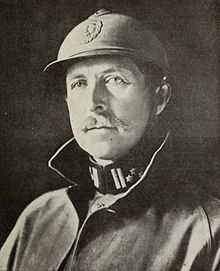 A man wearing a military uniform, and helmet, with a moustache.