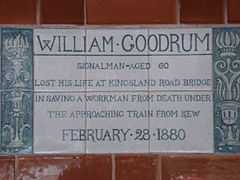 A tablet formed of six standard sized tiles, bordered by green flowers in the style of the Arts and Crafts movement. The tablet reads "William Goodrum signalman aged 60 lost his life at Kingsland road bridge in saving a workman from death under the approaching train from Kew February 28, 1880".