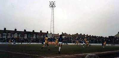 Two teams taking part in a football match, one in blue shirts and the other in yellow. Stands full of spectators are visible in the background.