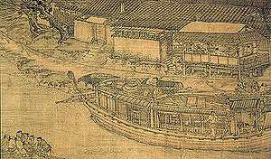 A close up view of a large trading barge crewed by multiple people. The barge has wooden walls surrounding it on all sides, and a thin line of tiled roof capping the walls, but not covering the interior of the ship. There are several windows built into the wall of the vessel.