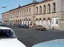 A white neo-Victorian station building, with a street full of parked 1970s cars in front of it.