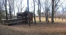 An open wooden structure with a ramp leading up to it is one the edge of a large open field, with large leafless trees around.