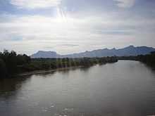  A wide flat river flows between tree-lined banks. Jagged hills rise in the distance.