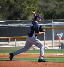 A man throwing a baseball wearing a navy-blue baseball jersey and cap and gray baseball pants