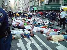 Street protest scene; people deliberately lying down on a busy city street, surrounded by onlookers and police