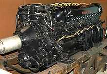 A front right view of a black-painted aero engine, the words 'Rolls-Royce' appear in red. The engine has yellow electrical wiring and is sitting on a wooden pallet.