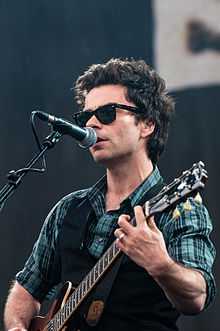 A man wearing sunglasses, a waistcoat and a checked shirt, singing into a microphone whilst playing an electric guitar