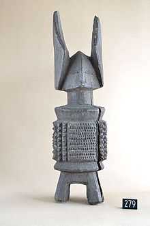 An image of a carved deity named Ikenga, the grey wooden piece has legs, a stylised but simple body, a trinagular head and shallow facial features and two horns around 1/3 it's size